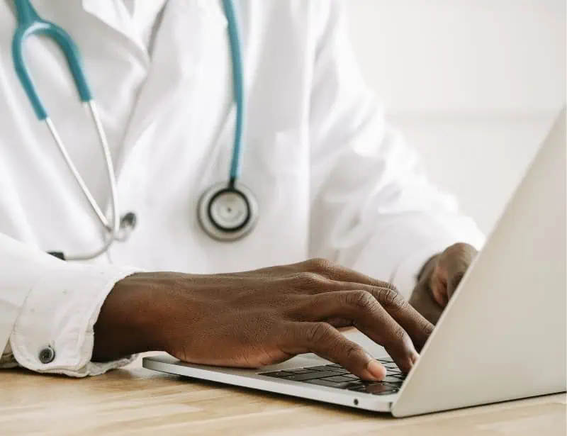 black doctor with blue stethoscope around neck typing on laptop