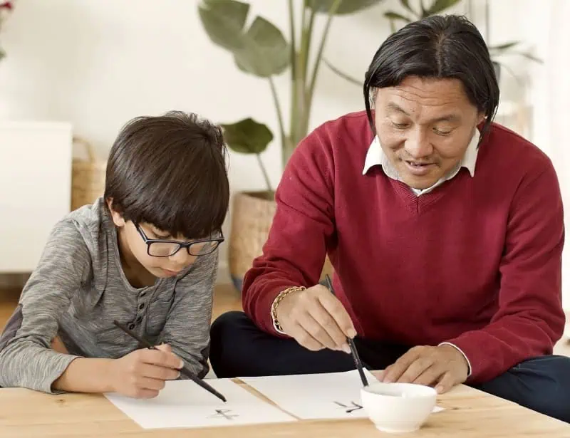 father with red top teaching son with glasses how to do Chinese calligraphy
