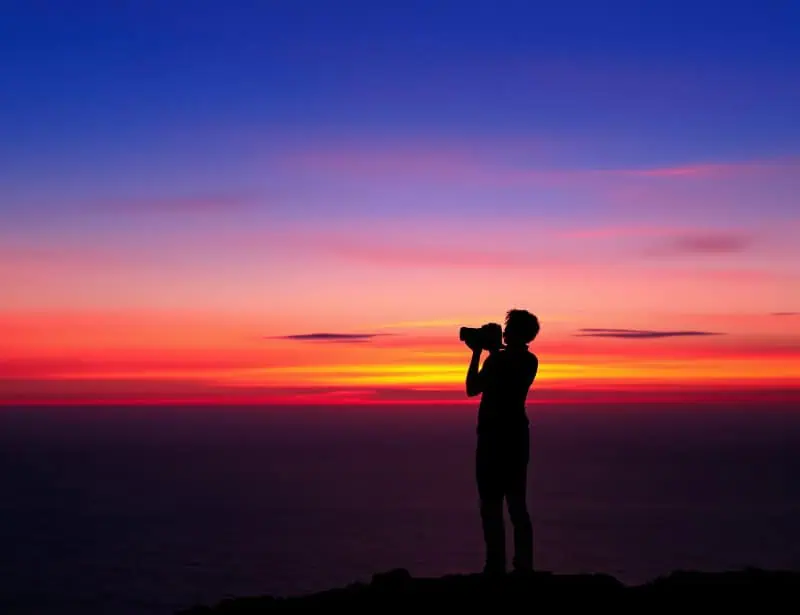 Silhouette of a photographer standing on rocks against a vibrant sunset over water, capturing the dramatic sky with their camera.