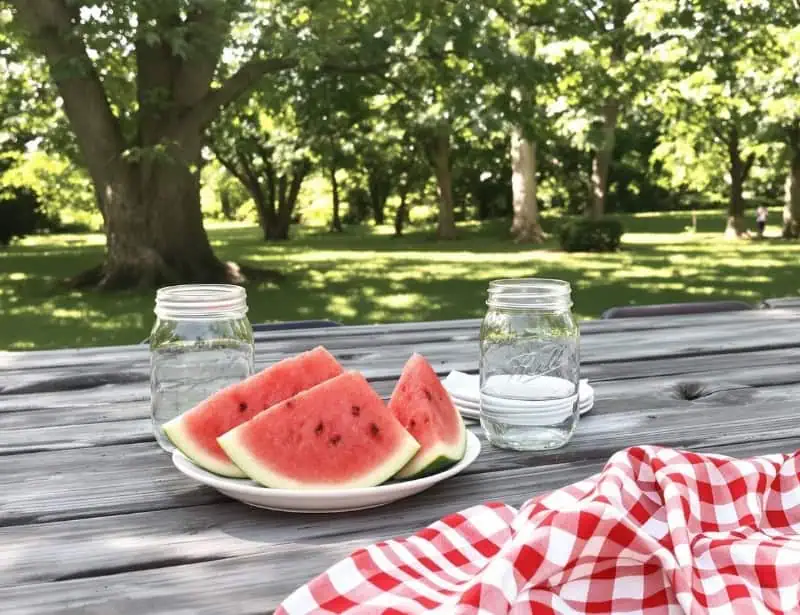 Weathered wooden picnic table with watermelon slices on a white plate, mason jar of water, and checkered cloth under dappled sunlight.