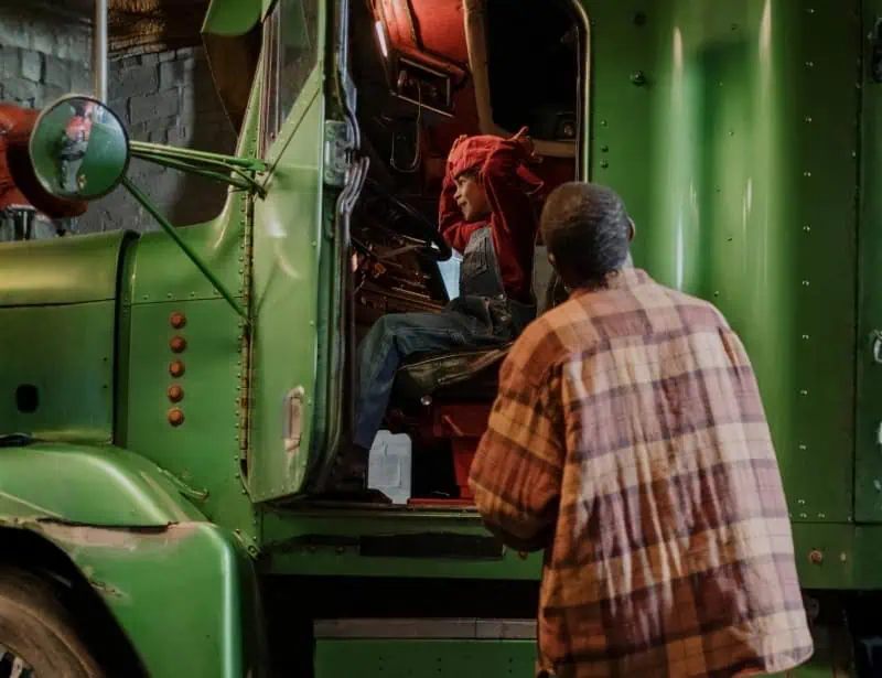 truck driver looking at boy wearing cap and smiling while on truck driving seat 