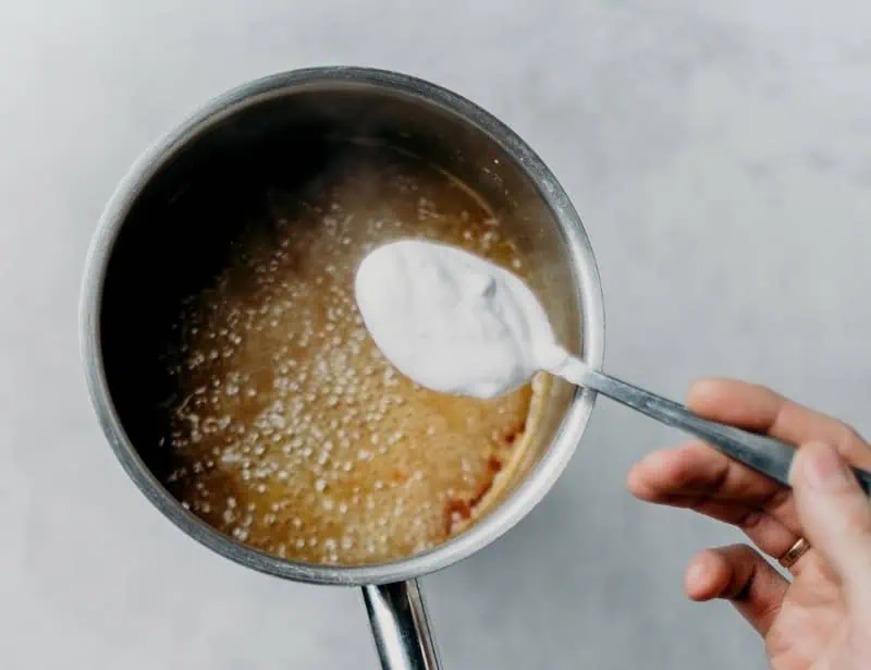 hand about to add one tablespoon of white powder on saucepan with caramelizing sugar