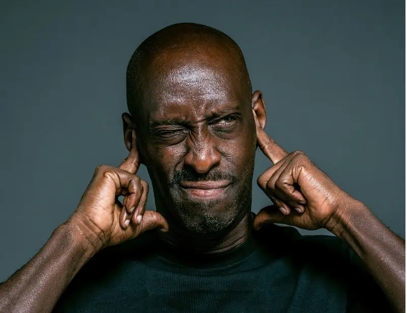 black bald man wearing black shirt visibly in pain while holding both ears
