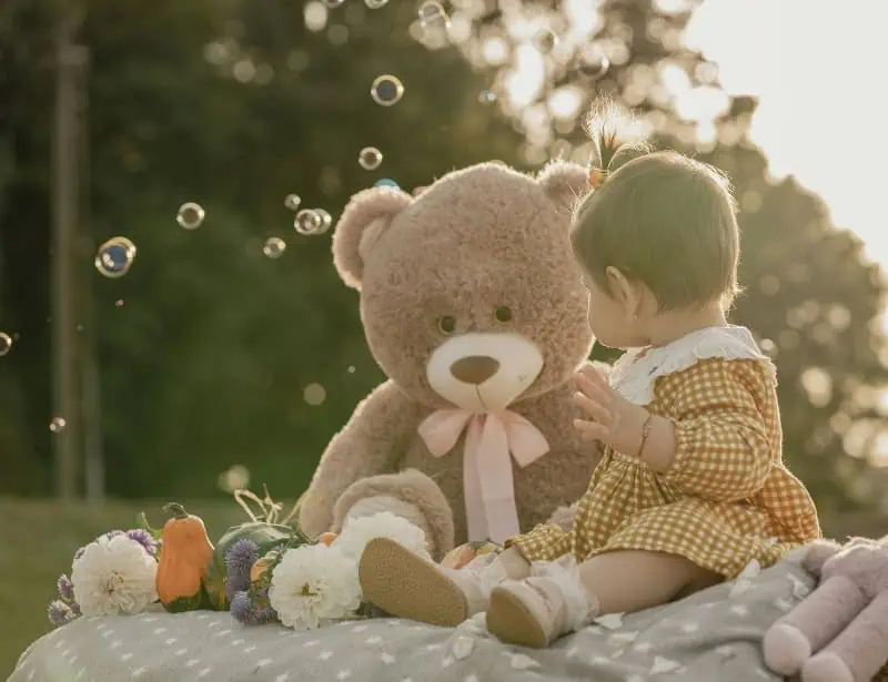 baby girl sitting on picnic blanket with brown teddy bear pink ribbon in park with bubbles