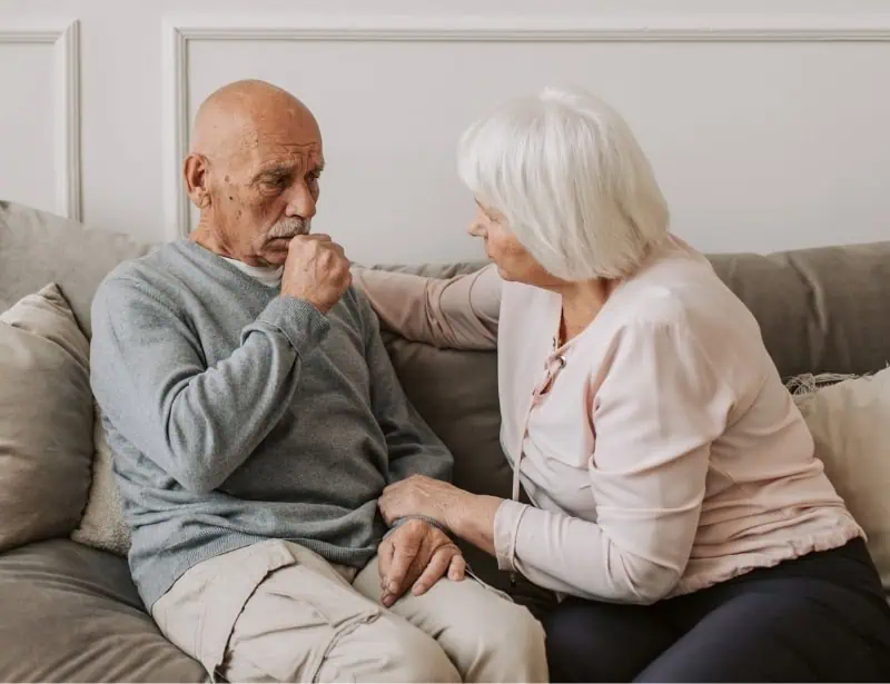 elder woman comforting coughing old man sitting on sofa