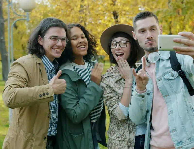 four adult friends smiling while taking selfie in park