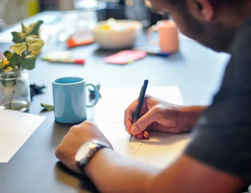 over the shoulder man writing on piece of paper on table with blue mug