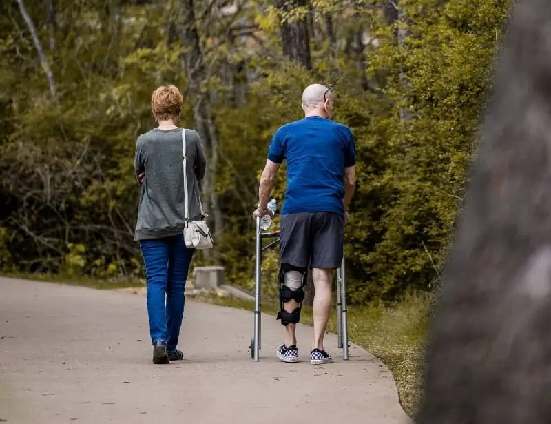 back of woman with crossbody bag and man with crutches walking on park path 