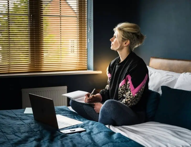 blonde middle aged woman looking at window with opened laptop notebook and pen while on bed