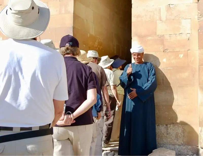 egyptian guide in blue robe showing thumbs up to tourists entering ancient structure