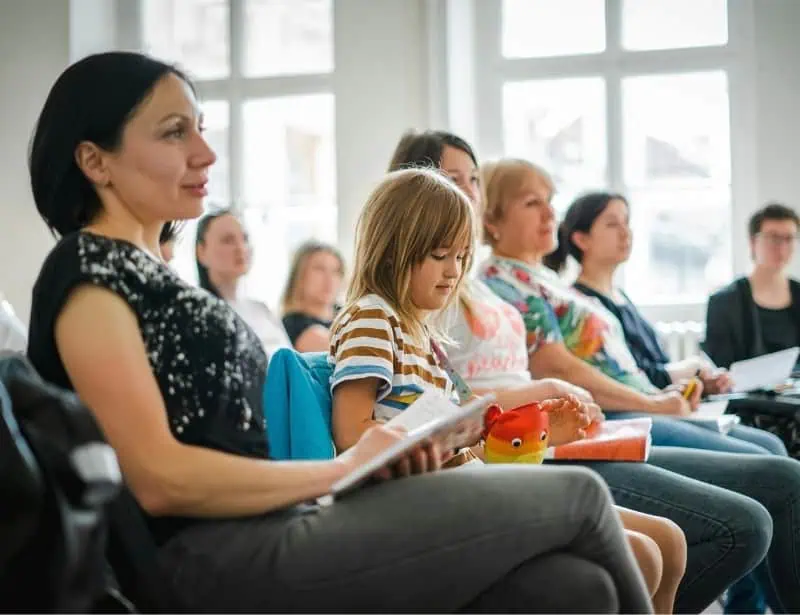 mother beside daughter listening at front with notebook together with other adults