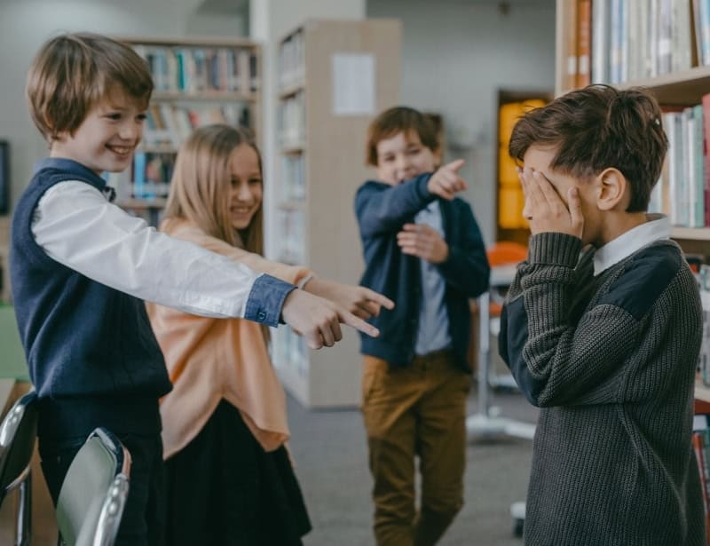 three middle school students laughing and pointing at child covering face with hand in library