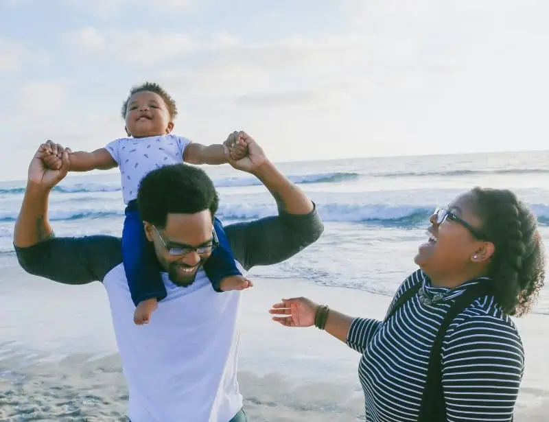 black american family father holding baby son on shoulders and mother laughing at the beach daytime
