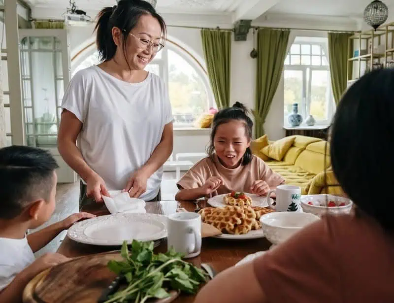 mother presenting plate of waffles on table with smiling children already sat