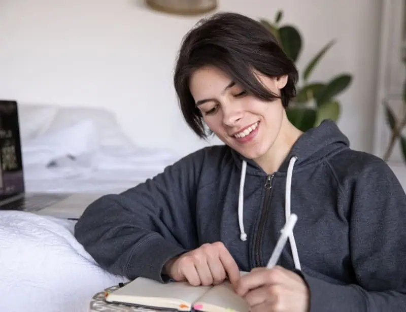 woman wearing dark gray hoodie smiling while writing something on notebook